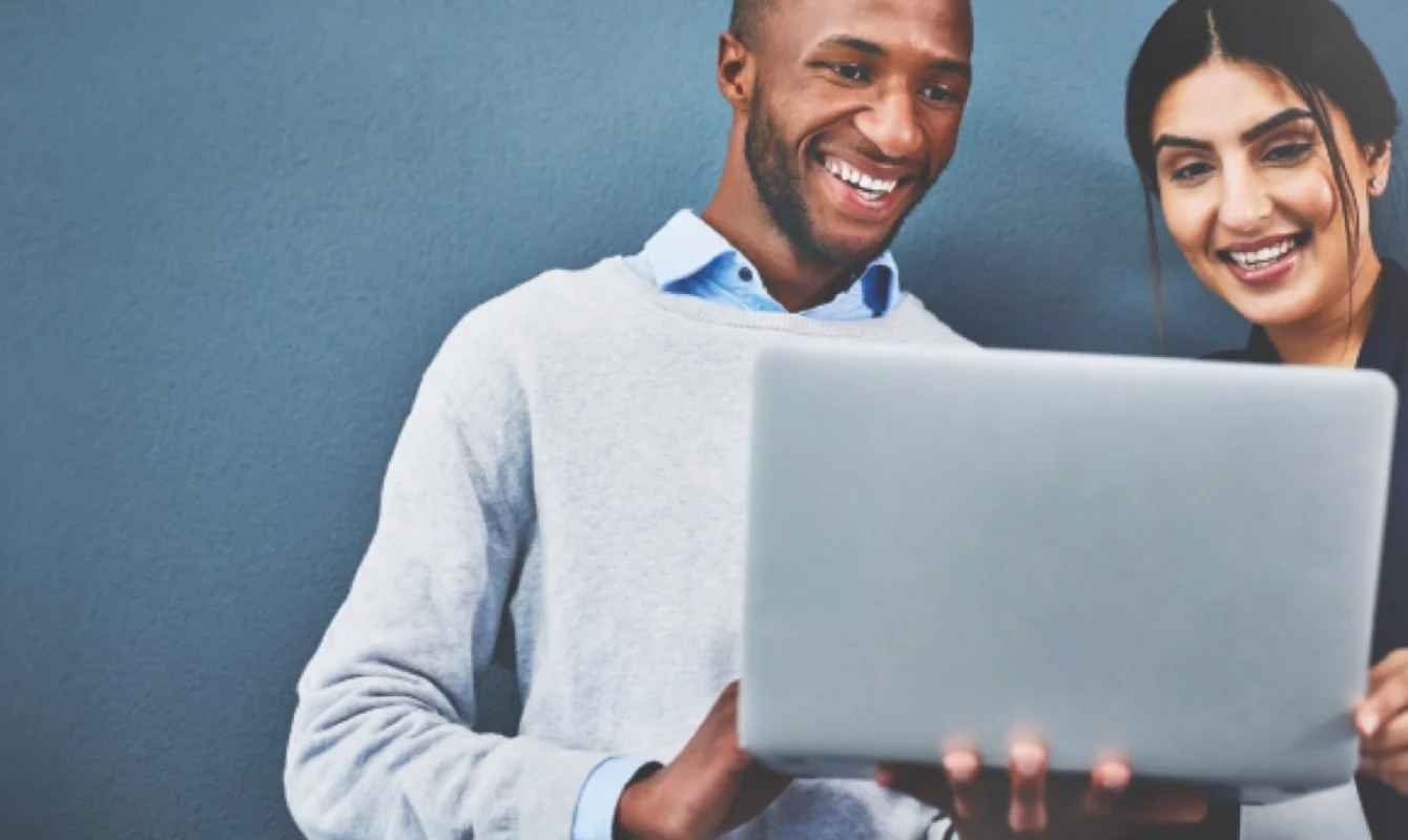 Two people smiling while looking at a laptop screen against a grey wall background.